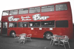 red double decker bus converted into a bar with side serving hatch. Metal tables and chairs set up in front of bus bar.