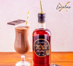 Image of Lucelas chocolate and Rum brown spirit in a bottle with a spirit pourer top, next to a large glass of chocolate cocktail with a gold and white straw and two squares of chocolate balanced on the side.