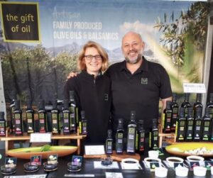 image of man and lady smiling at camera standing behind gift of oil exhibitor stand selling a range of oils and balsamic dressings, several sample bowls open at front of display.