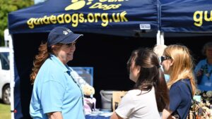 image of fundraising woman wearing cap who is standing next to a guide dogs gazebo, talking to two women that look interested in the cause.
