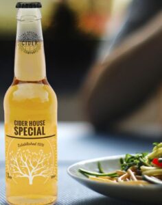 image of light coloured cider in 330ml sized bottle label reads cider hoouse special established in 1976, image of apple tree on bottle. the bottle is on a table next to a bowl of noodles and vegetables.