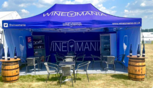 image of dark blue Wineomania 6x3 meter open gazeba with three seperate seating areas consisting of a metal table with three metal chairs for wine tasting , two fridges containing many bottles of wine and two tables made from barrells on either side of the gazebo-each with three bottles of red wine on them.