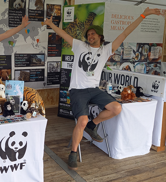 image of man sitting down with arms open in WWF gazebo, he is sitting between two tables which have several soft toy pandas on them and wildlife leaflets.