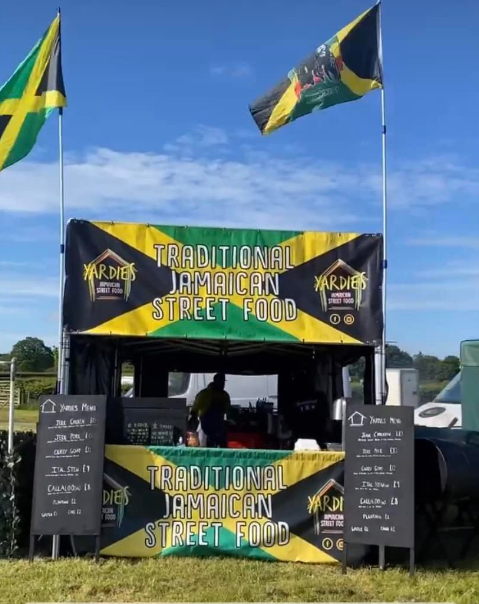 Jamaican street food gazebo, with two big Jamaican flags flying either side of the gazebo. two blackboard menus on each side of the gazebo . Jamaican flag above and below the serving hatch with street food stall name Yardies written on it.