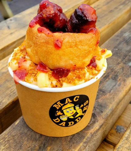 mac and cheese is served in brown card cup, fgenerous portion topped with Yorkshire pudding and cranberry. serving is on a wooden table.