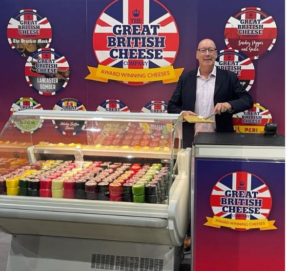 cheese stand consisting of large refrigerator filled with various truckle cheeses, background of stand has great British cheese logos placed on board. man in front of sign serving a piece of cheese to sample.