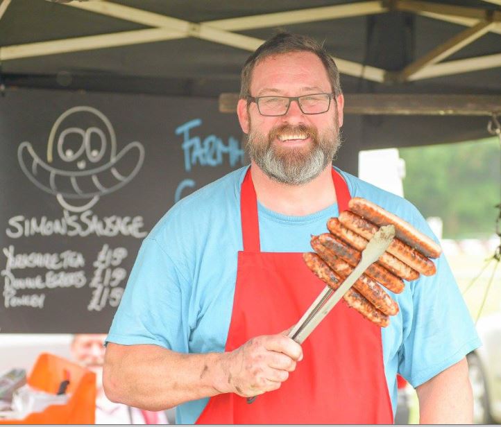 image of man underneath gazebo, wearing blue t-shirt and red apron, holding up 6 bratwurst style sausages in bbq tongs