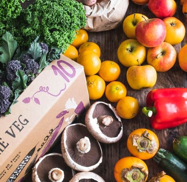 image of fruit and veg delivery box with lots of fresh veg and fruit spilling out of it including cut field mushrooms, red peppers, tangerines, apples and leafy green vegetables.