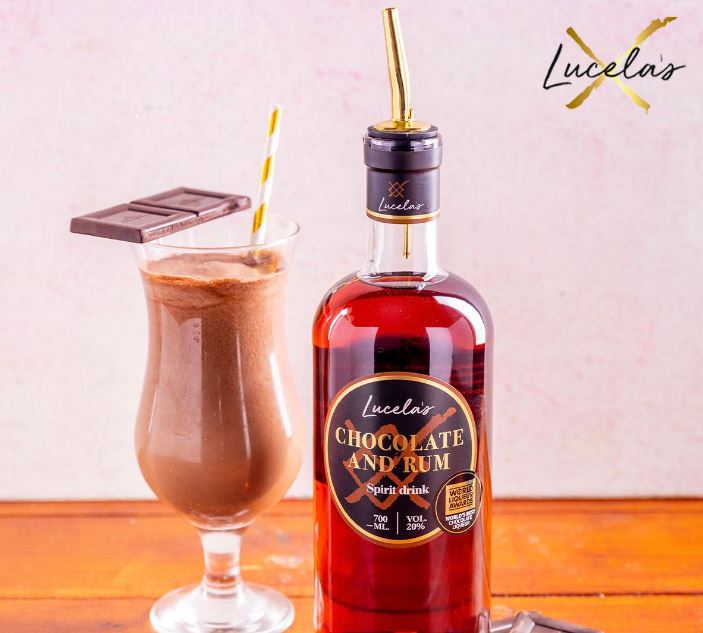 Image of Lucelas chocolate and Rum brown spirit in a bottle with a spirit pourer top, next to a large glass of chocolate cocktail with a gold and white straw and two squares of chocolate balanced on the side.