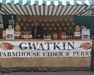 image of open market stand , with counter area across the front baring various bottles of cider. man standing behind counter. many cider flagon bottles hanging up on wall behind man. sign at front of counter reads, Gwatkin Farmhouse Cider and Perry in large black text. green and white striped canopy style roof on market stand.