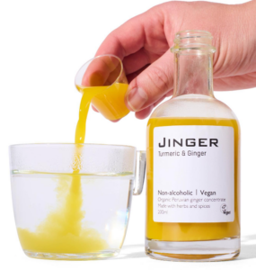 image of hand pouring shot of orange liquid into clear glass. glass bottle in front of hand which contains nearly full bottle of orange liquid. label reads Jinger Turmeric and Ginger non Alcoholic.