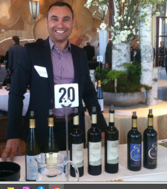 image of smiling man in checked shirt and jacket standing behind a table containing 7 bottles of open red wine set out for wine tasting.