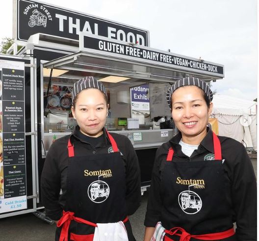 Two Thai ladies dressed in black and red somtam branded aprons smiling in front of somtam thai food truck.