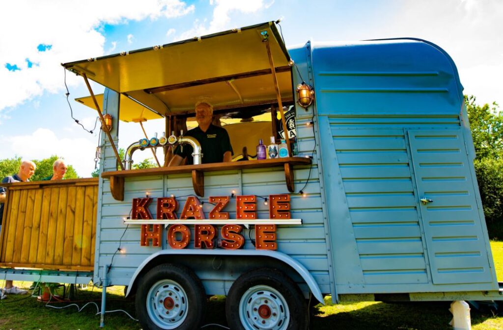 image of mobile bar, blue converted horsebox with side hatcgh. sign beaneath hatch reads Krazee Horse in red stand out letters, photo credit to Rich Nelson Photography