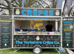 Image of yorkeshire crepe mobile street food trailer with opening sides and serving hatch. m,nu is written on the side doors, neon sign saying crepes inside the trailer. lots of serving sauces on outside serving ledge such as nutella, honey, syrup.