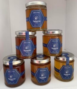 image of 6 jars of hedgerow honey stacked in a pyramid formation, with three jars on the bottom, two in the middle and one on top. the jars all contain a dark amber honey with a gold lid and a blue label.