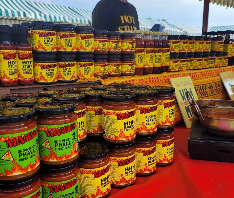 image of many jars of spiced sauces , chutney and curry blends set up on a exhibitors table with red table cloth. man behind stand wearing a hot stuff tshirt.