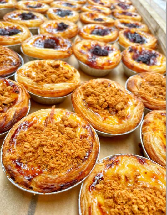 Image of several Portuguese tarts laid out on a counter, several different toppings including biscoff , black cherry and original.