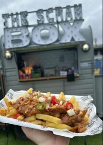 Image of the Scran box catering trailer with a portionn of loaded fries being held up in front of the trailer.