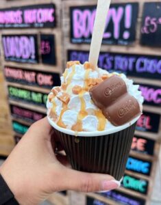 image of decadent looking hot chocolate in hand, in disposable cup, lots of whipped cream caramel sauce and galaxy caramel chocolate pieces.